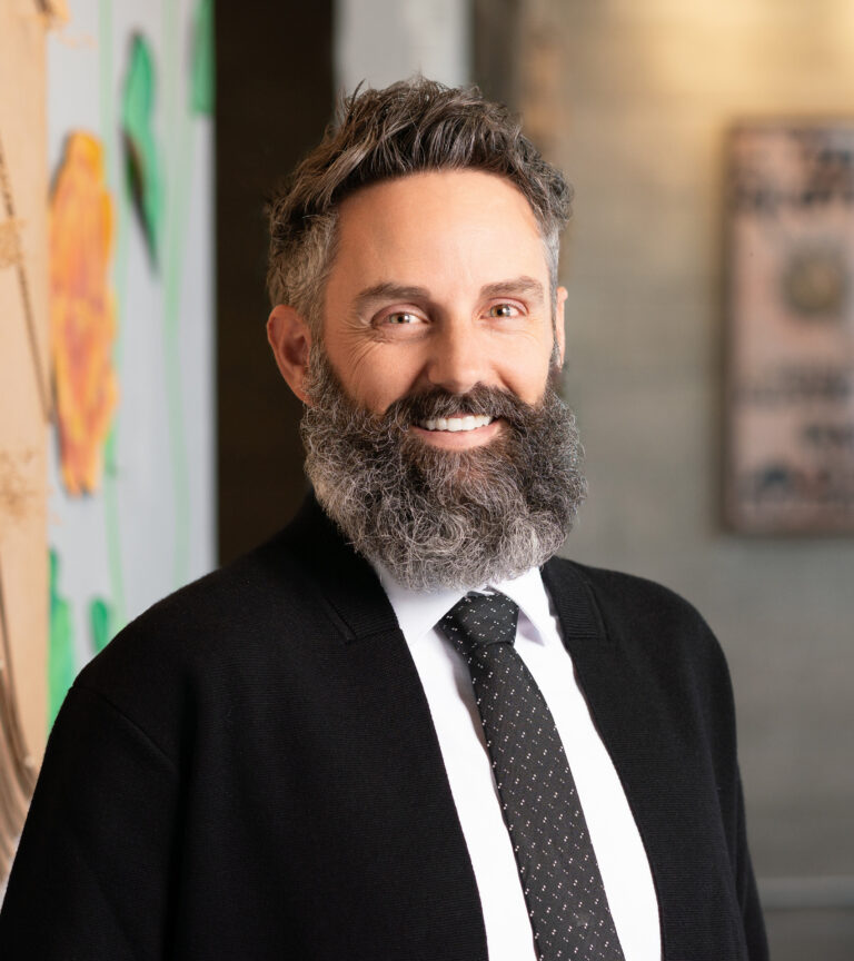 Smiling bearded man in a black cardigan, white shirt, and tie, standing indoors against a softly blurred background