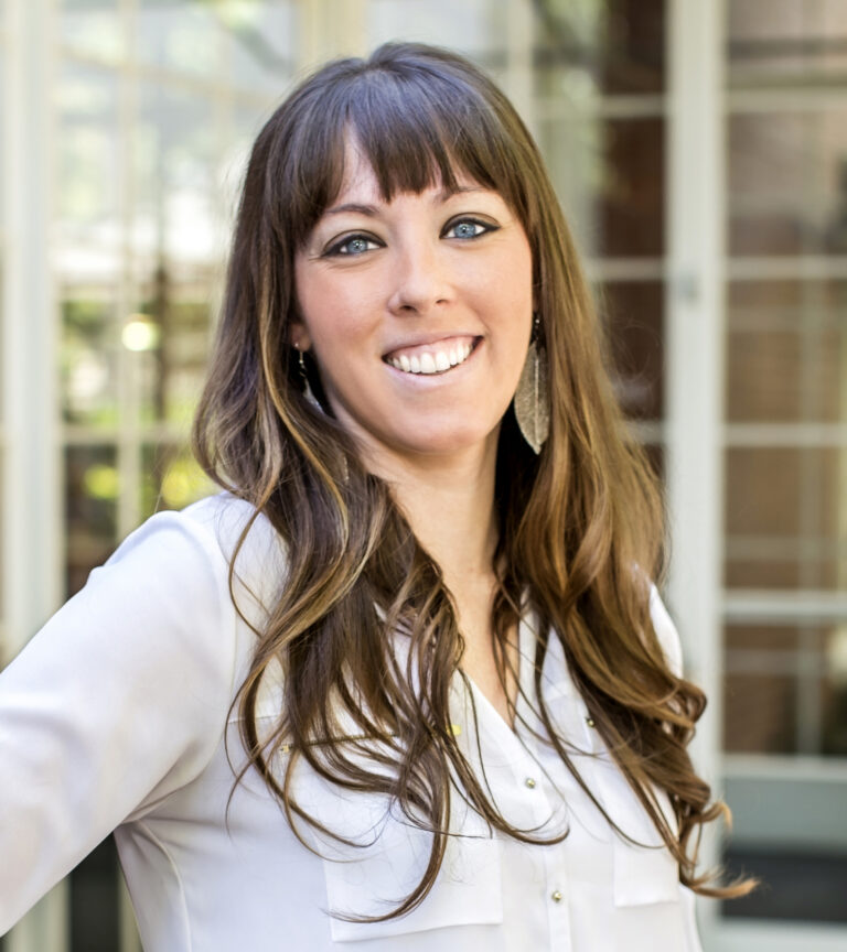 Smiling woman with long brown hair and bangs wearing a white blouse, standing indoors near large windows