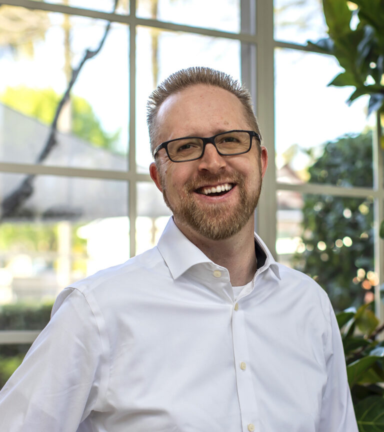 Smiling man with glasses and a beard in a white shirt, standing indoors by a sunlit window and plants