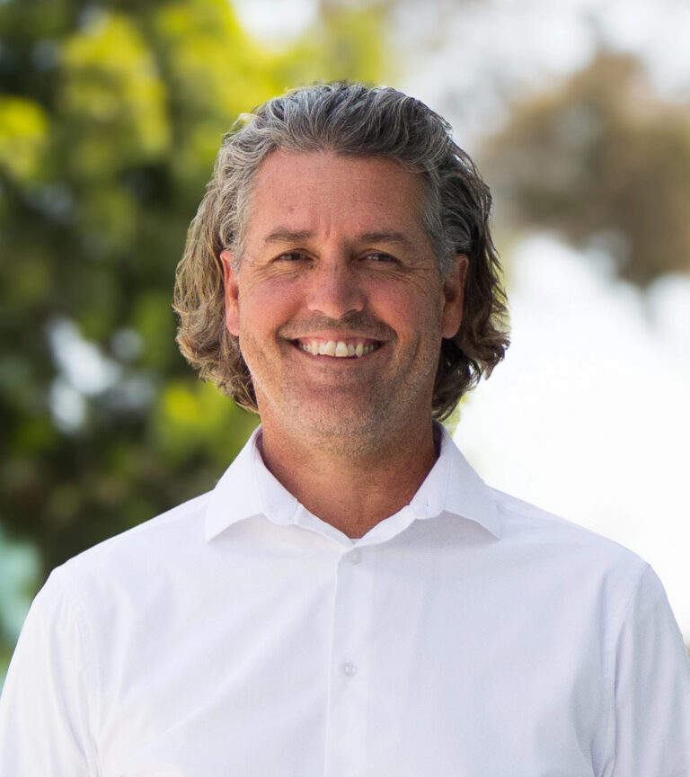 Smiling man with wavy gray hair in a white button-down shirt outdoors against a blurred leafy background