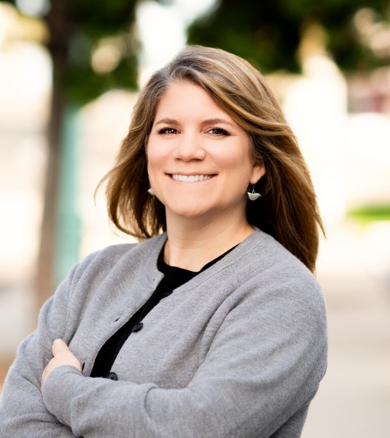 Smiling woman in a gray cardigan and earrings, posed outdoors with a blurred background