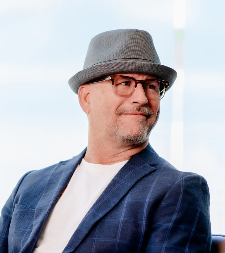 Smiling man in glasses and a gray hat wearing a blue plaid blazer, looking to the side indoors