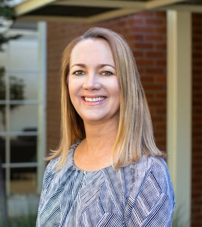 Smiling blonde woman in a patterned blouse standing outdoors in front of a brick building