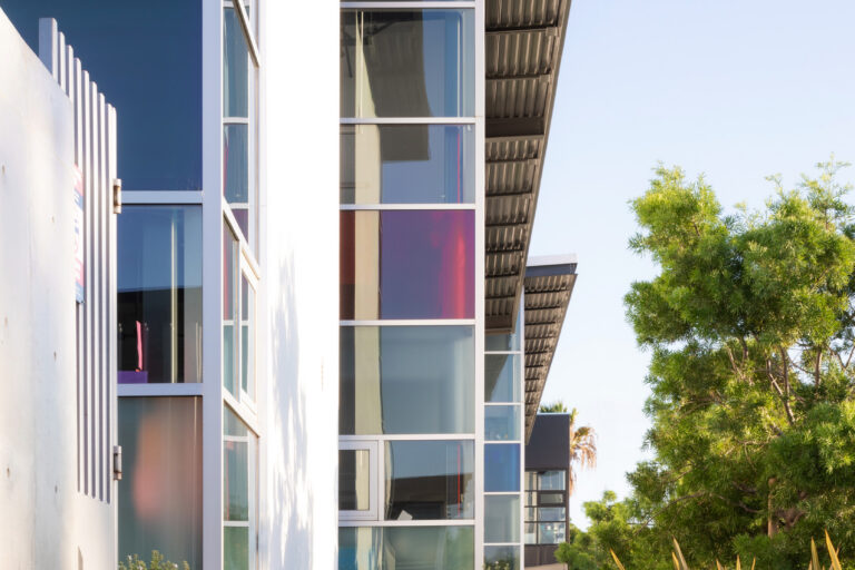 Modern apartment building with tall glass windows, metal awnings, and a landscaped walkway beside trees and shrubs
