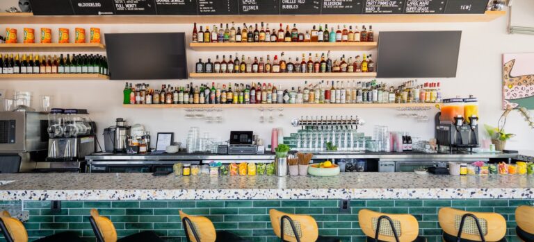 Modern bar interior with green-tiled counter, row of stools, liquor shelves, and a large drink menu board.