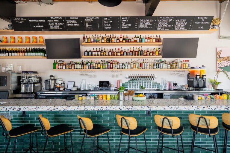 Modern bar interior with green-tiled counter, row of stools, liquor shelves, and a large drink menu board.
