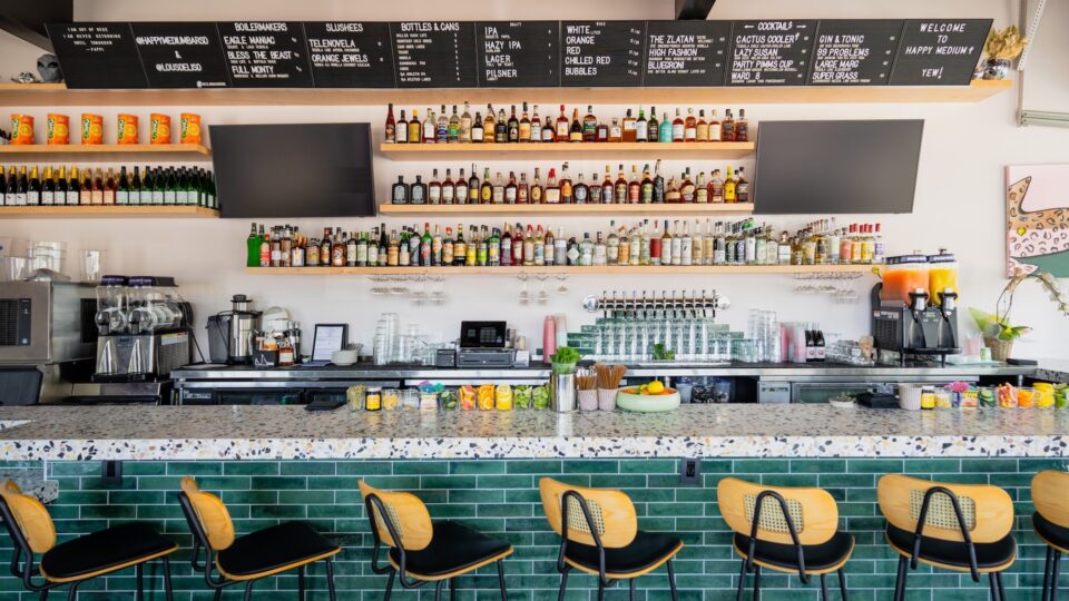 Modern bar interior with green-tiled counter, row of stools, liquor shelves, and a large drink menu board.