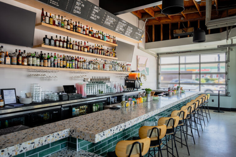 Modern bar interior with terrazzo counter, tall stools, liquor shelves, beer taps, and menu boards by a garage-style window