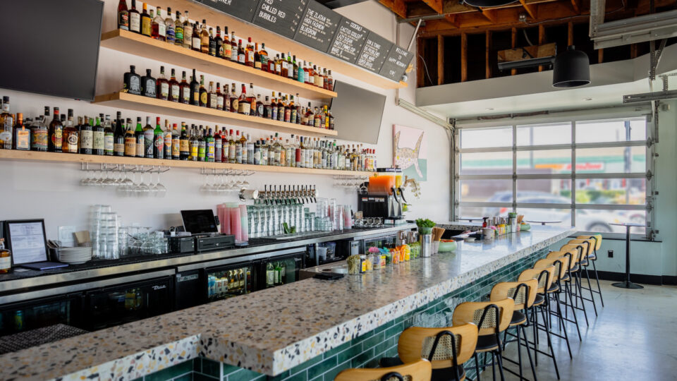 Modern bar interior with terrazzo counter, tall stools, liquor shelves, beer taps, and menu boards by a garage-style window