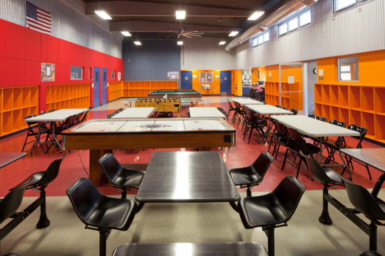 Bright recreation room with tables, orange cubbies, an air hockey table, foosball table, pool tables, and an American flag.