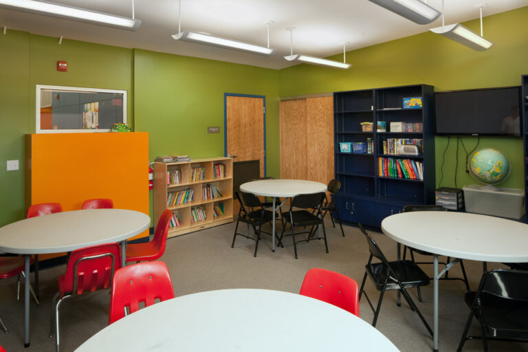 Children’s classroom with round tables, red chairs, bookshelves, a TV, and a globe against green walls