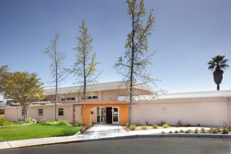 Front entrance of the James A. Bronner Family Branch building with trees, lawn, and a clear blue sky.