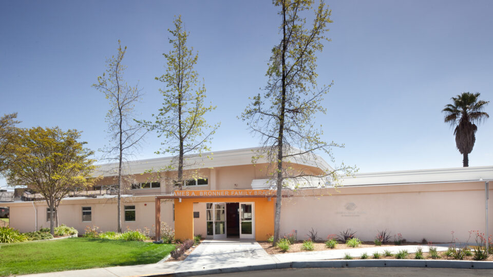 Front entrance of the James A. Bronner Family Branch building with trees, lawn, and a clear blue sky.