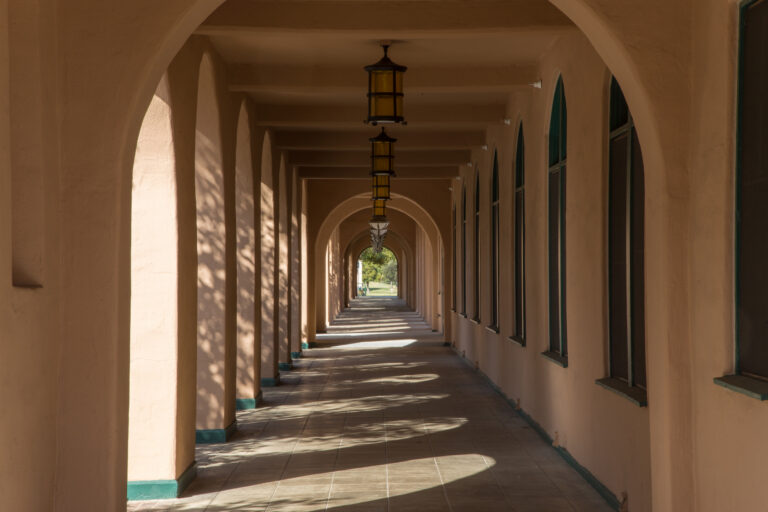 Sunlit arched corridor with hanging lanterns, tiled floor, and repeating shadows leading to a distant garden path