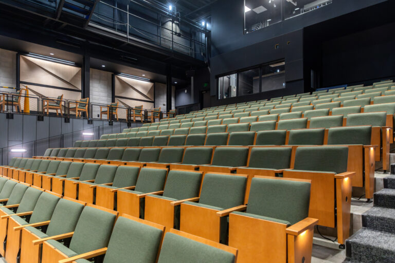 Modern auditorium with tiered rows of green seats, side steps, and an upper balcony with lounge seating