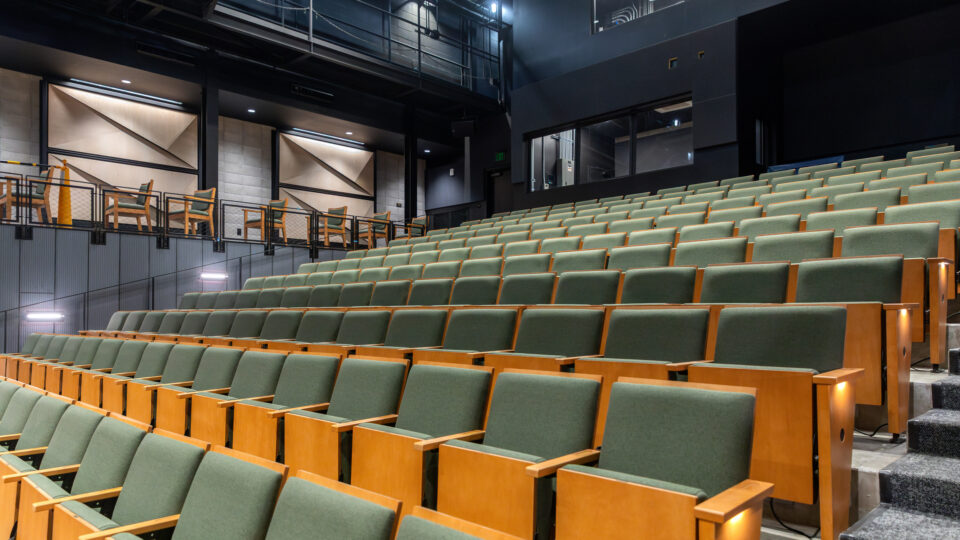 Modern auditorium with tiered rows of green seats, side steps, and an upper balcony with lounge seating