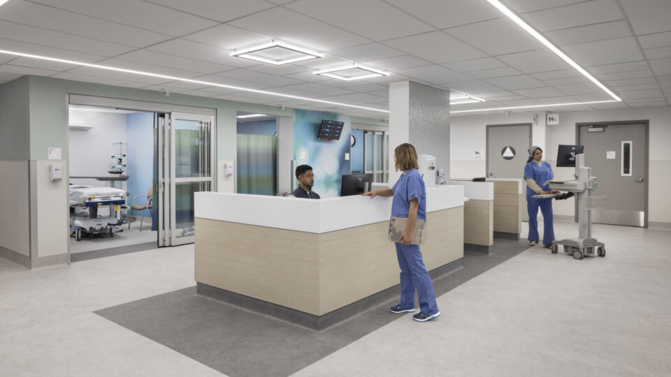 Modern hospital nurse station with staff in blue scrubs, patient rooms, and medical equipment in a bright hallway