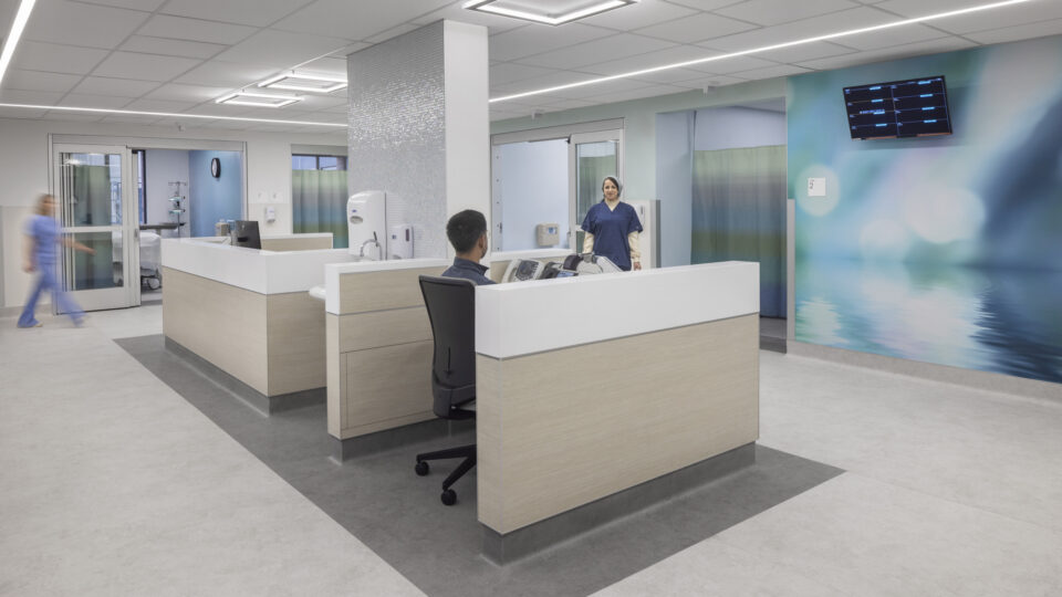 Modern hospital nurses’ station with staff at desks and in hallways, surrounded by patient rooms and soft blue wall graphics