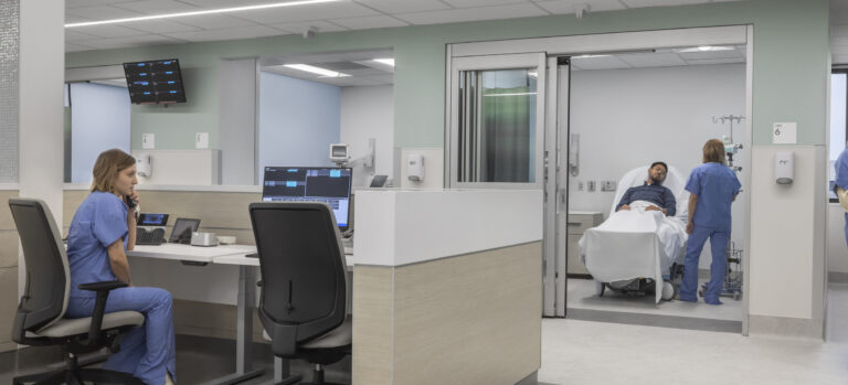 Modern hospital nurses' station with staff at computers and a patient in a bed being attended in an exam room