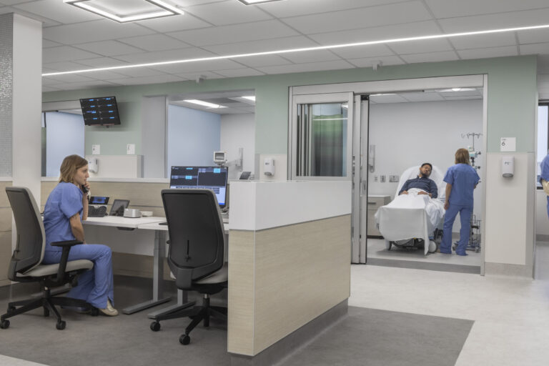 Modern hospital nurses' station with staff at computers and a patient in a bed being attended in an exam room