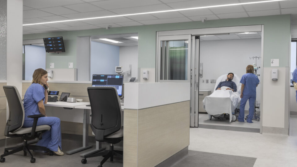 Modern hospital nurses' station with staff at computers and a patient in a bed being attended in an exam room