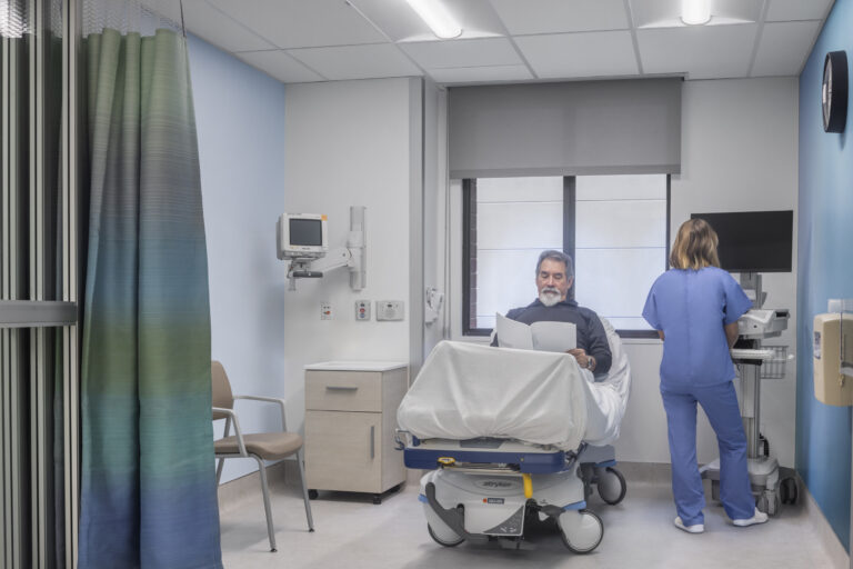 Man sitting on a hospital bed reading papers while a nurse in blue scrubs works at a computer in the room.