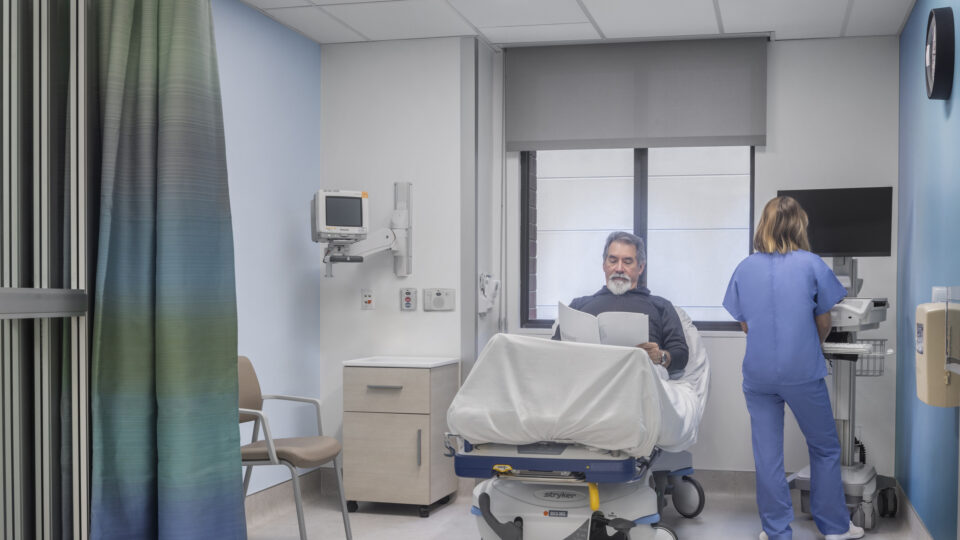 Man sitting on a hospital bed reading papers while a nurse in blue scrubs works at a computer in the room.