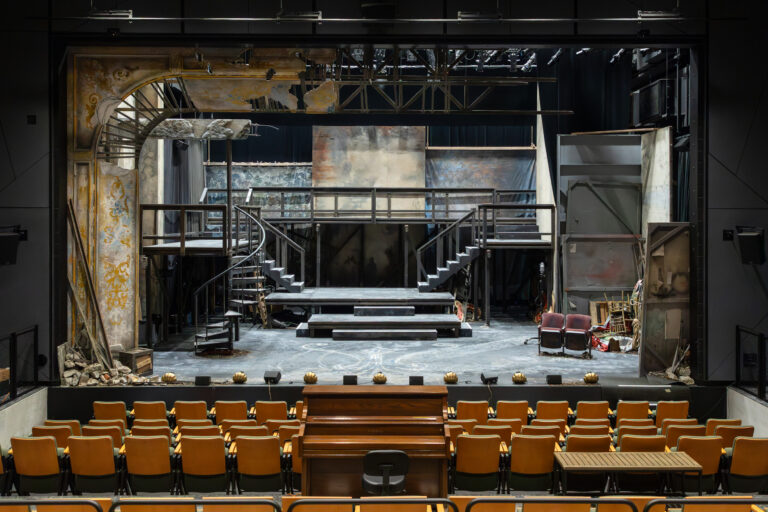 Theater stage set with worn staircases, platforms, debris, and empty audience seats viewed from the auditorium.