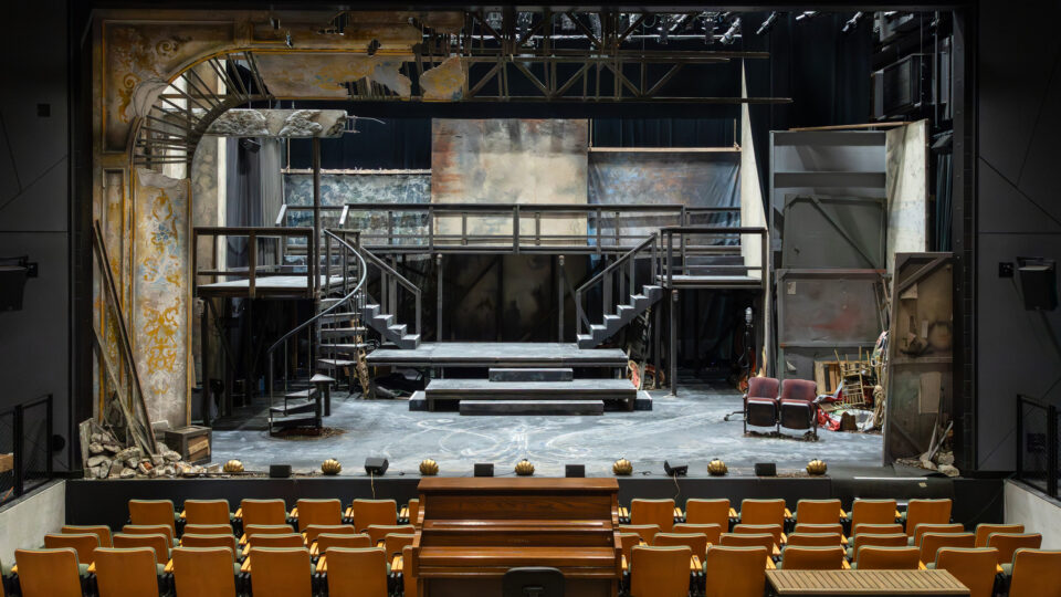 Theater stage set with worn staircases, platforms, debris, and empty audience seats viewed from the auditorium.