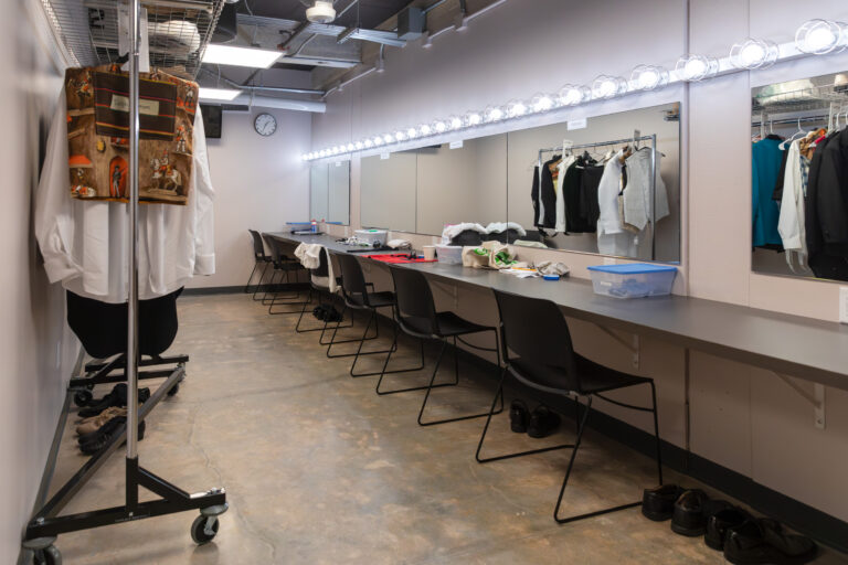 Backstage dressing room with lighted mirrors, chairs, costume racks, and shoes lined up on the floor
