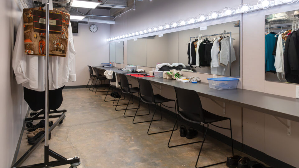 Backstage dressing room with lighted mirrors, chairs, costume racks, and shoes lined up on the floor