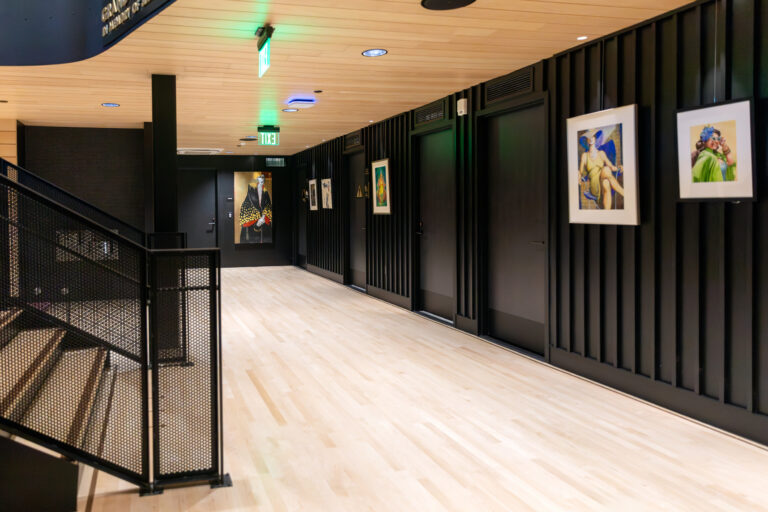 Modern hallway with black paneled walls, light wood floor, framed artwork, and a stair railing under EXIT signs