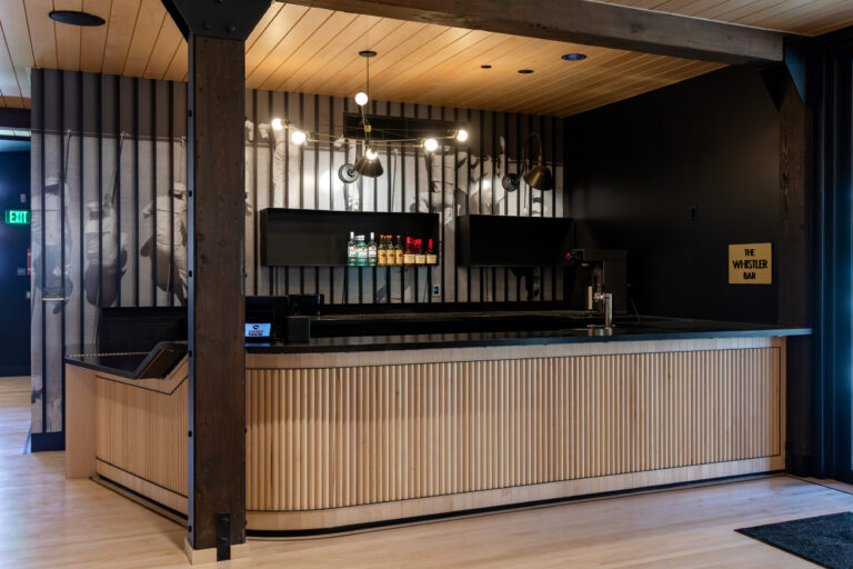 Modern black-and-wood bar with bottle display, wall mural, and a sign reading The Whistler Bar