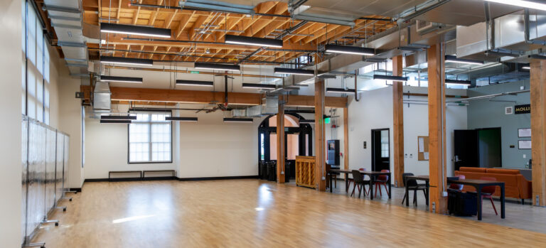 Spacious modern hall with wood floors, exposed beams and ductwork, hanging lights, and tables and seating along one side