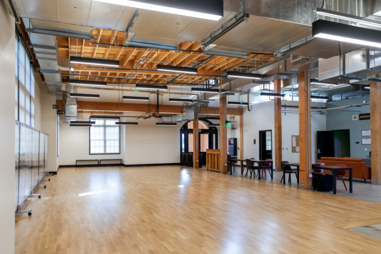Spacious modern hall with wood floors, exposed beams and ductwork, hanging lights, and tables and seating along one side