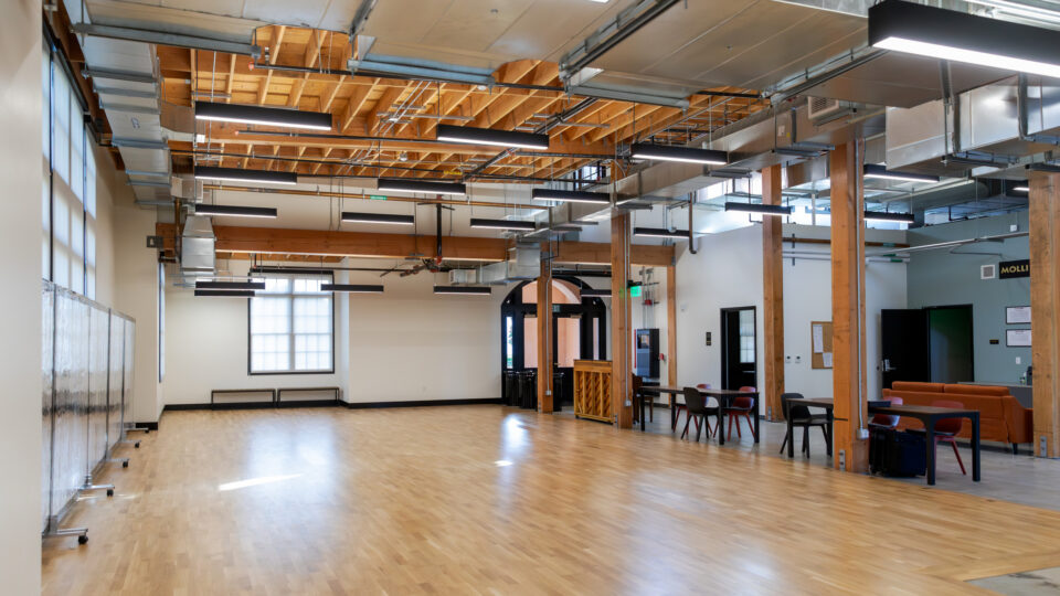 Spacious modern hall with wood floors, exposed beams and ductwork, hanging lights, and tables and seating along one side