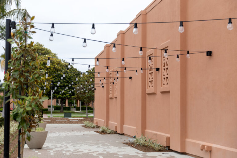 String lights hanging above a paved walkway beside a peach-colored building with trees and planters.