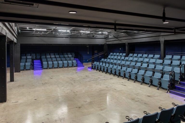 Empty black-box theater with tiered blue seating, illuminated aisle steps, and an open floor performance area