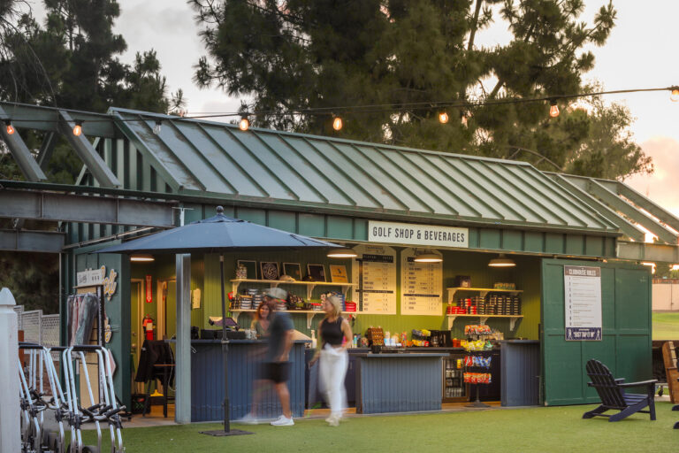 Golf shop and beverages stand at dusk, with string lights, golf push carts, and blurred people walking by