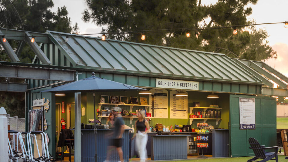 Golf shop and beverages stand at dusk, with string lights, golf push carts, and blurred people walking by
