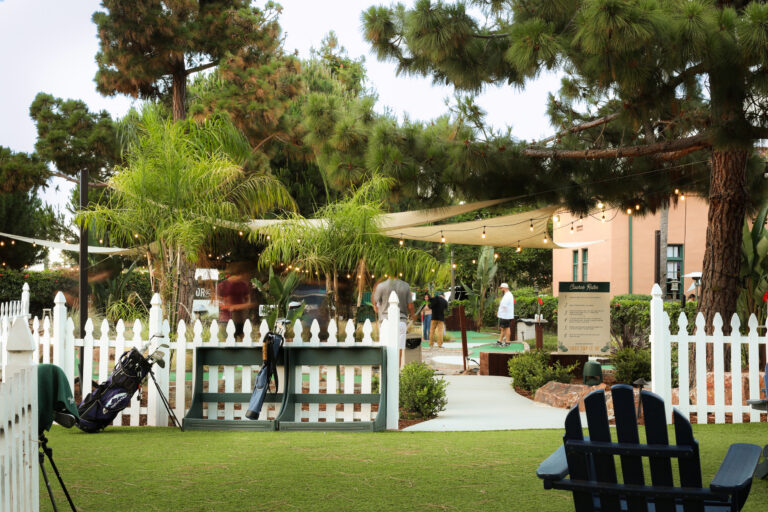 Mini golf course with palm trees, string lights, and golfers beyond a white picket fence.