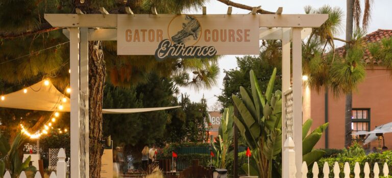 Entrance to a mini golf course with a Gator Course sign, white pergola, tropical plants, and string lights.