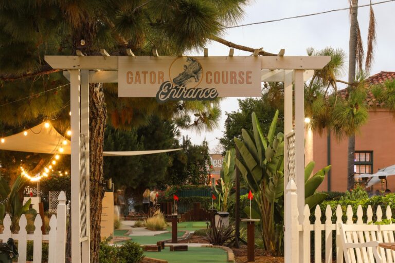 Entrance to a mini golf course with a Gator Course sign, white pergola, tropical plants, and string lights.