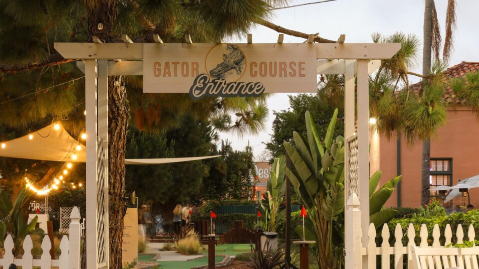 Entrance to a mini golf course with a Gator Course sign, white pergola, tropical plants, and string lights.