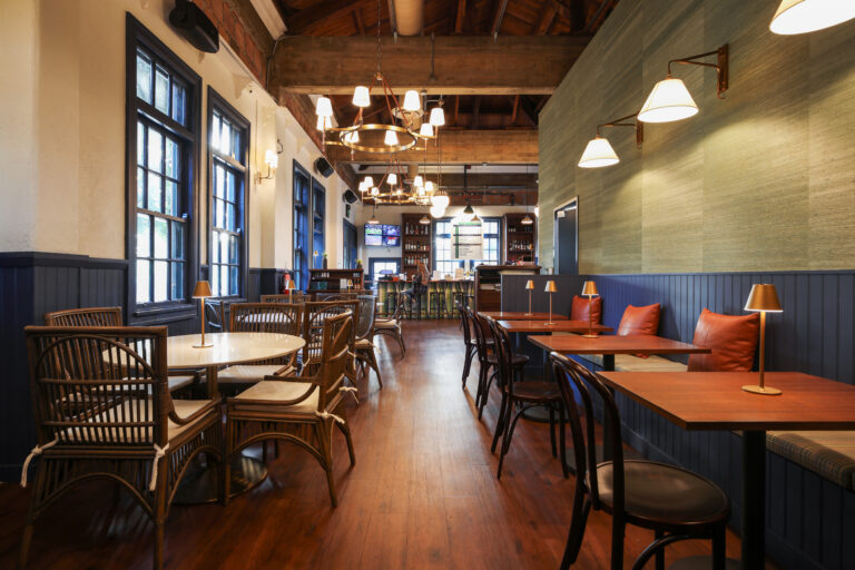 Warm, stylish restaurant interior with wooden floors, wicker chairs, booth seating, tall windows, and hanging lights.
