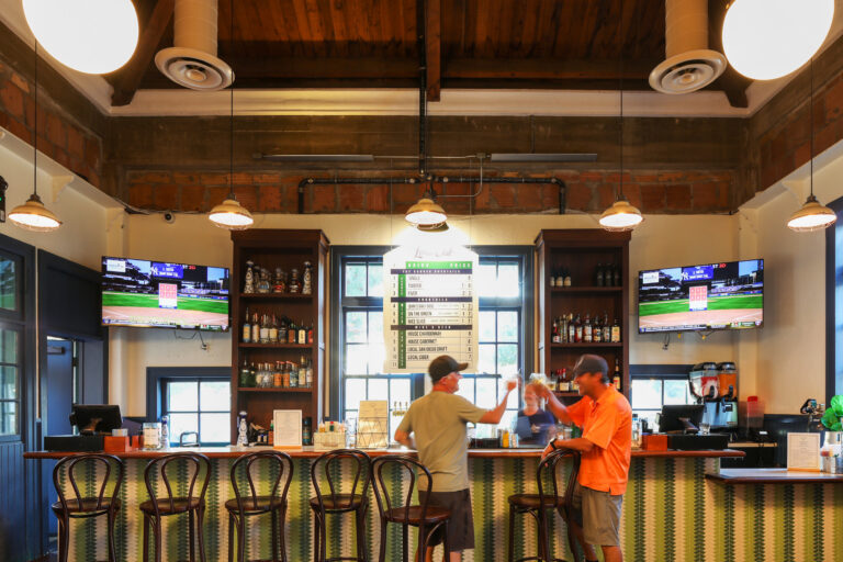 Two men talking at a modern bar with TV screens, high stools, and exposed wood beams overhead.