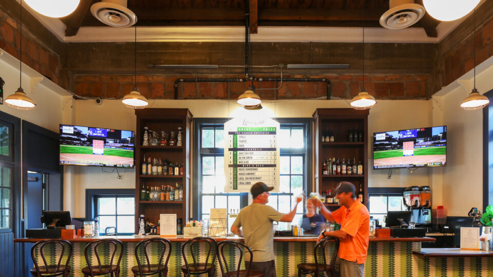 Two men talking at a modern bar with TV screens, high stools, and exposed wood beams overhead.