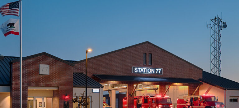 Brick fire station labeled Station 77 at dusk, with fire engines parked outside and U.S. and California flags flying.
