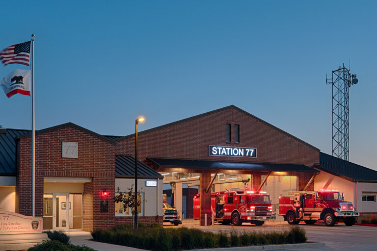 Brick fire station labeled Station 77 at dusk, with fire engines parked outside and U.S. and California flags flying.
