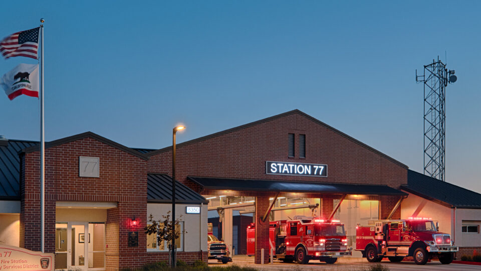 Brick fire station labeled Station 77 at dusk, with fire engines parked outside and U.S. and California flags flying.